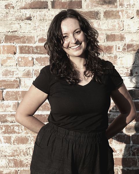 Julia Comstock-Ross, smiling, standing against a sunlit brick wall
